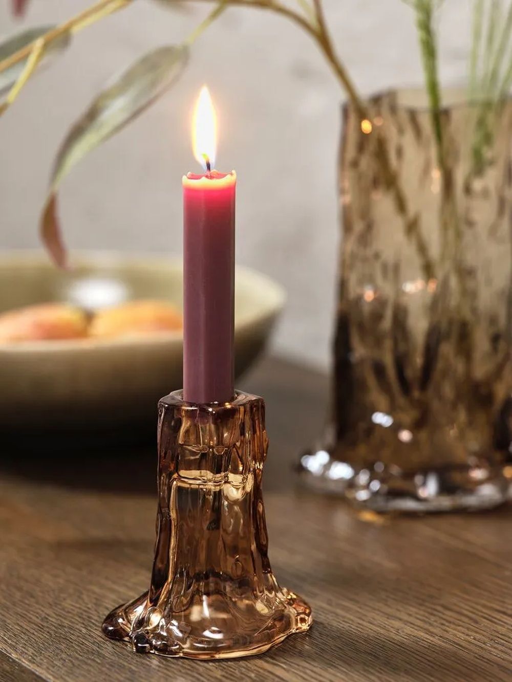 Vela de color burdeos encendida sobre un portavelas de cristal junto a un jarrón decorativo.