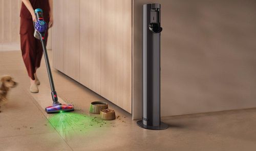 Persona utilizando una aspiradora Dyson V10 Konical para limpiar el suelo junto a un perro y varios restos de comida.