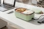 Fiambrera eléctrica con comida caliente sobre una mesa de oficina junto a un ordenador portátil