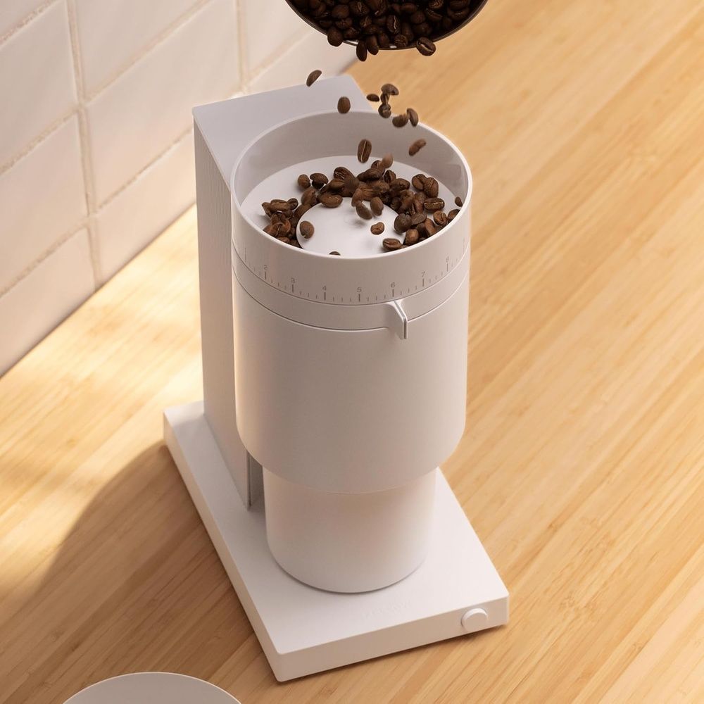 Granos de café cayendo en el depósito de un molinillo eléctrico blanco sobre una encimera.