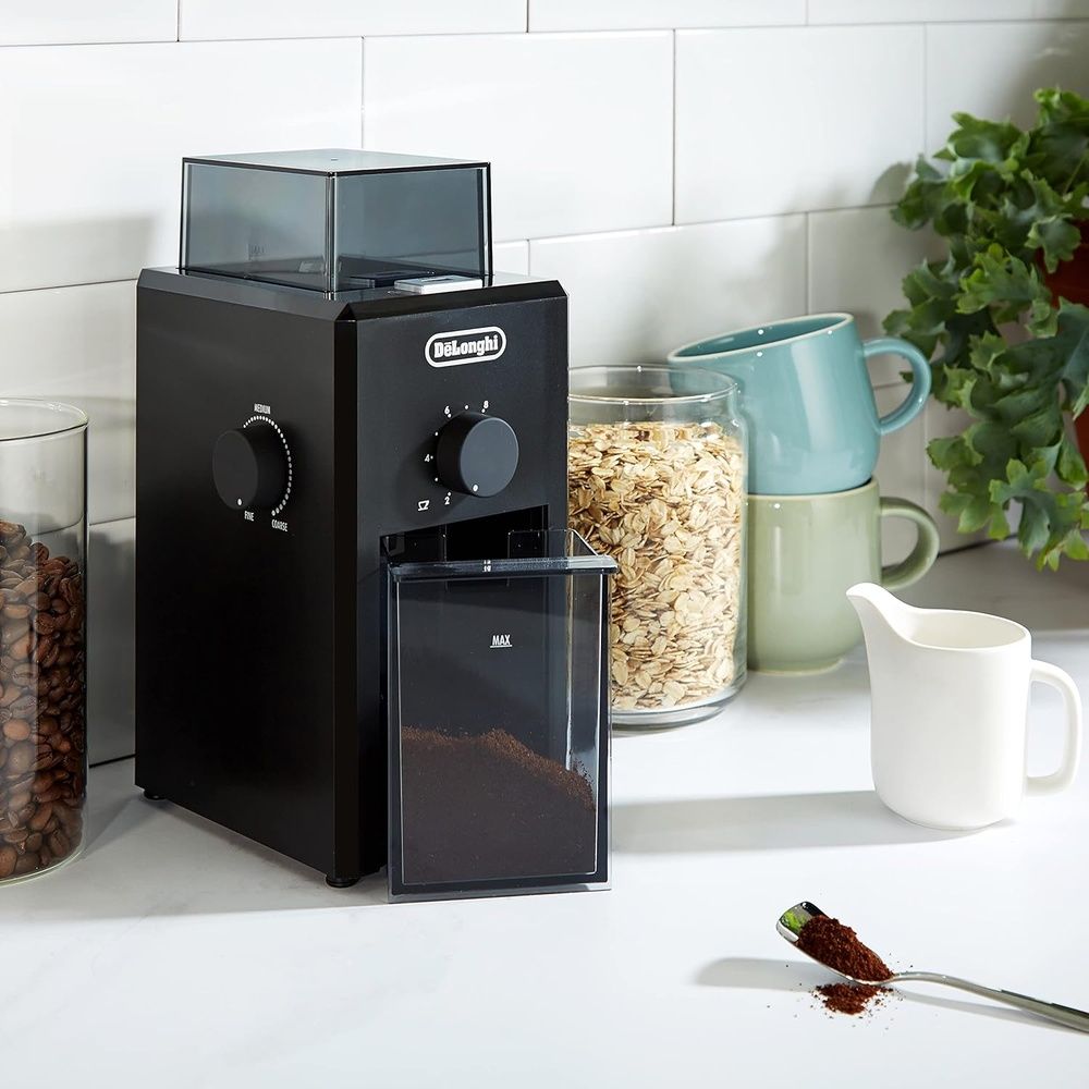 Molinillo de café eléctrico De'Longhi junto a granos de café y varias tazas en una cocina.