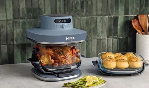 Freidora de aire Ninja CRISPi PRO XL con recipiente de cristal cocinando un pollo junto a otro recipiente con pan.