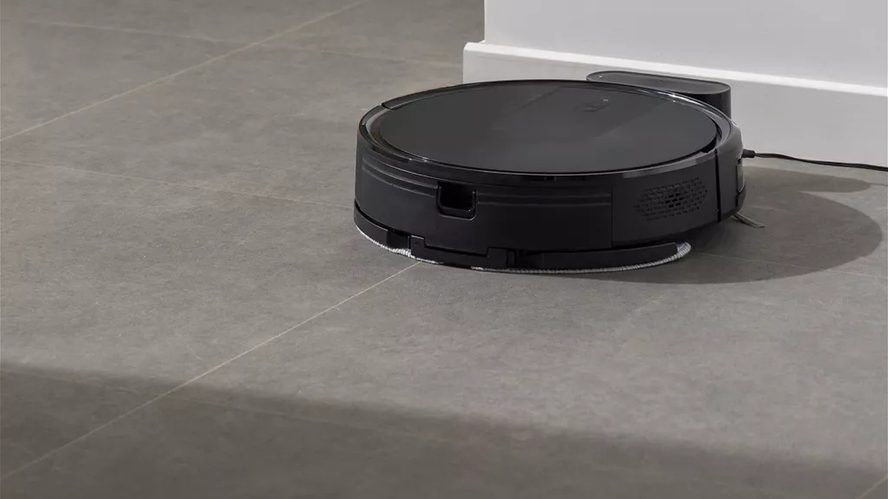 Robot aspirador negro en funcionamiento sobre el suelo de una vivienda, cerca de una pared y su base de carga