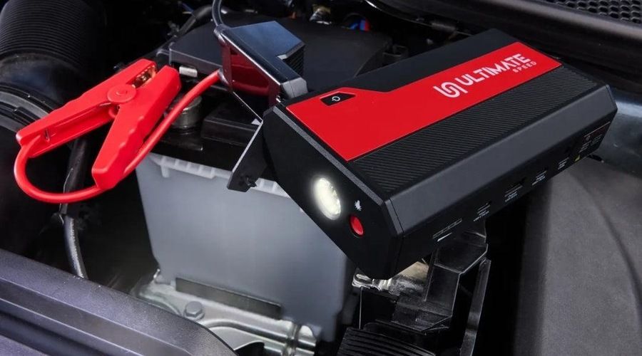 Primer plano del arrancador portátil Ultimate Speed conectado a la batería de un coche mediante sus pinzas, con la linterna LED encendida.