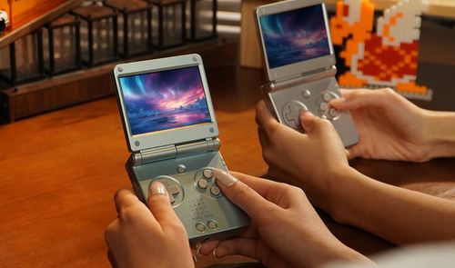 Dos personas sostienen consolas Anbernic RG SP abiertas sobre una mesa de madera, con videojuegos al fondo.
