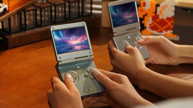 Dos personas sostienen consolas Anbernic RG SP abiertas sobre una mesa de madera, con videojuegos al fondo.