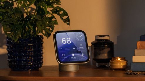 Apple HomeView sobre un mueble de madera, junto a una planta y varios objetos decorativos, con controles del hogar en pantalla.