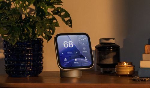 Apple HomeView sobre un mueble de madera, junto a una planta y varios objetos decorativos, con controles del hogar en pantalla.