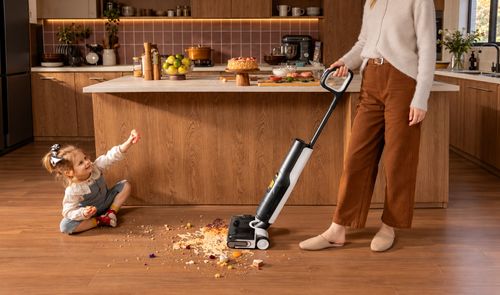 Mujer limpiando restos de comida con una aspiradora fregadora en una cocina, junto a una niña sentada en el suelo.