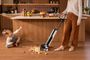 Mujer limpiando restos de comida con una aspiradora fregadora en una cocina, junto a una niña sentada en el suelo.