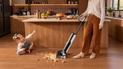 Mujer limpiando restos de comida con una aspiradora fregadora en una cocina, junto a una niña sentada en el suelo.
