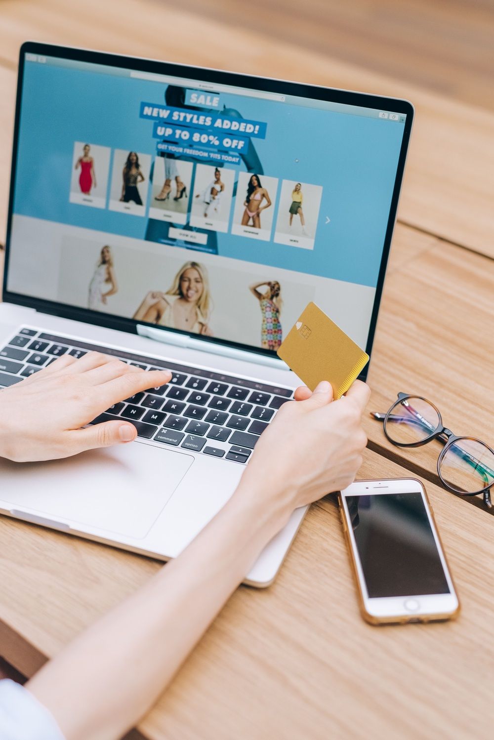 ersona consultando una tienda online en un portátil mientras sostiene una tarjeta bancaria, junto a un móvil y unas gafas.