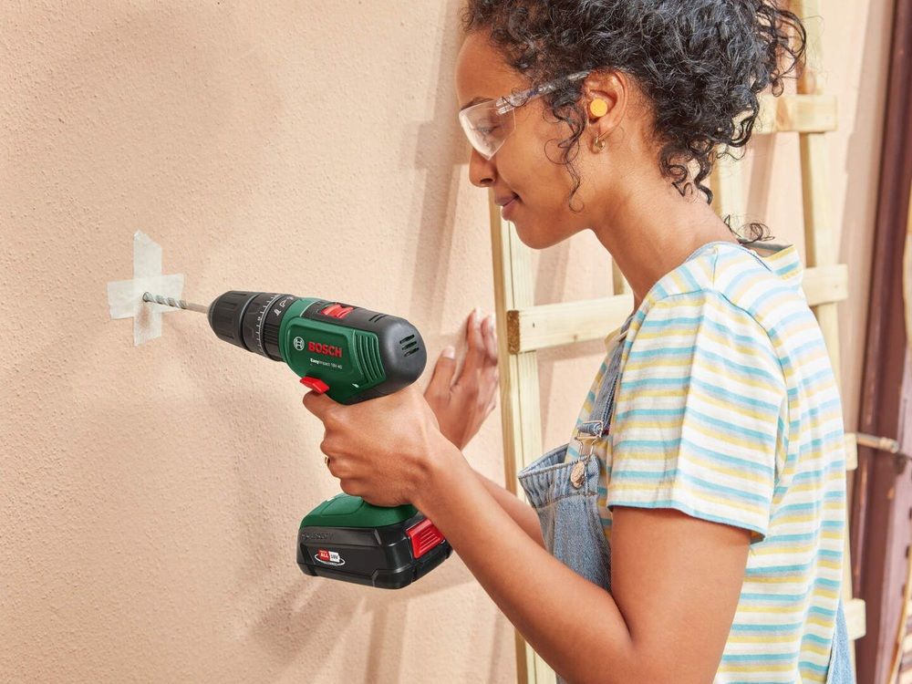 Mujer con gafas de protección taladrando una pared con un Bosch EasyImpact 18V-40 sobre una marca de cinta adhesiva.