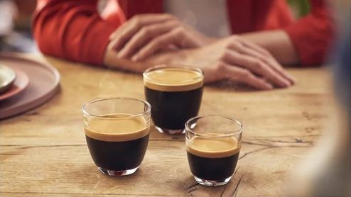 Tres vasos de café con una capa de crema sobre una mesa de madera, con una persona al fondo.