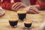 Tres vasos de café con una capa de crema sobre una mesa de madera, con una persona al fondo.