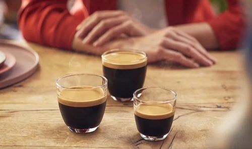 Tres vasos de café con una capa de crema sobre una mesa de madera, con una persona al fondo.