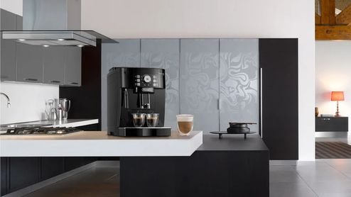 Cafetera automática De’Longhi negra sobre una isla de cocina, con dos cafés y un vaso de café con leche.