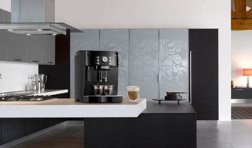 Cafetera automática De’Longhi negra sobre una isla de cocina, con dos cafés y un vaso de café con leche.