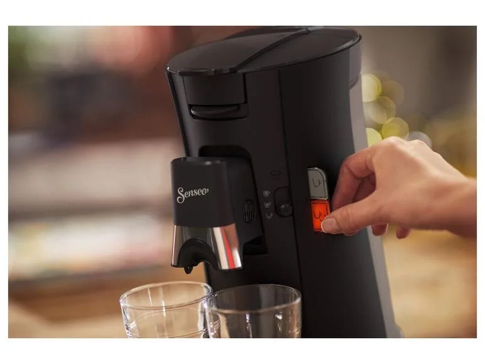 Una persona pulsa el botón de una cafetera Philips Senseo negra con dos vasos bajo la salida de café.