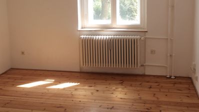 Radiador blanco bajo una ventana en una habitación con suelo de madera y luz natural.