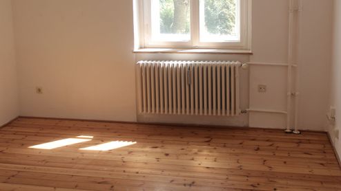 Radiador blanco bajo una ventana en una habitación con suelo de madera y luz natural.