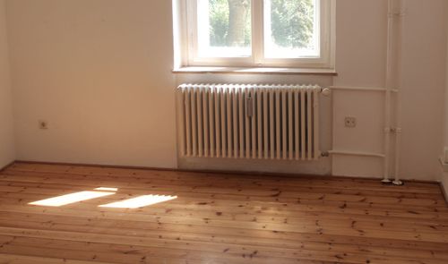 Radiador blanco bajo una ventana en una habitación con suelo de madera y luz natural.
