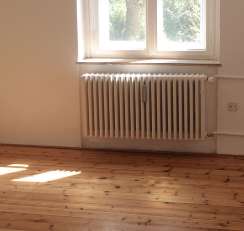 Radiador blanco bajo una ventana en una habitación con suelo de madera y luz natural.