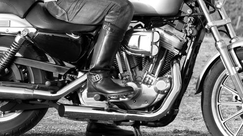 Bota alta de cuero negro apoyada en el reposapiés de una motocicleta, en una fotografía en blanco y negro.