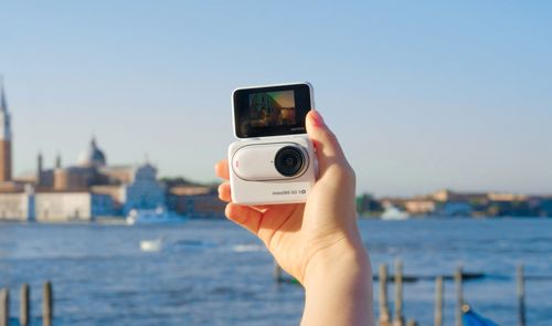 Insta360 GO 3S blanca sostenida en una mano, con la pantalla desplegada y Venecia de fondo.