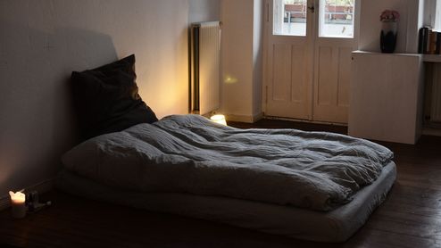 Colchón sobre suelo de madera con ropa de cama gris, junto a una ventana en un dormitorio.