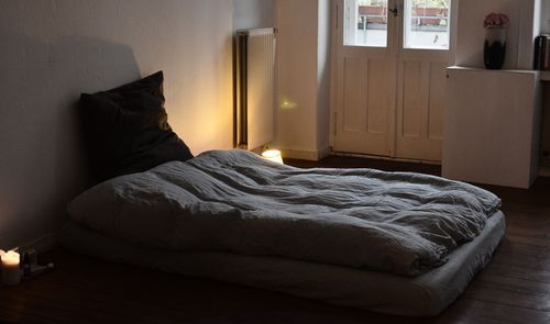 Colchón sobre suelo de madera con ropa de cama gris, junto a una ventana en un dormitorio.