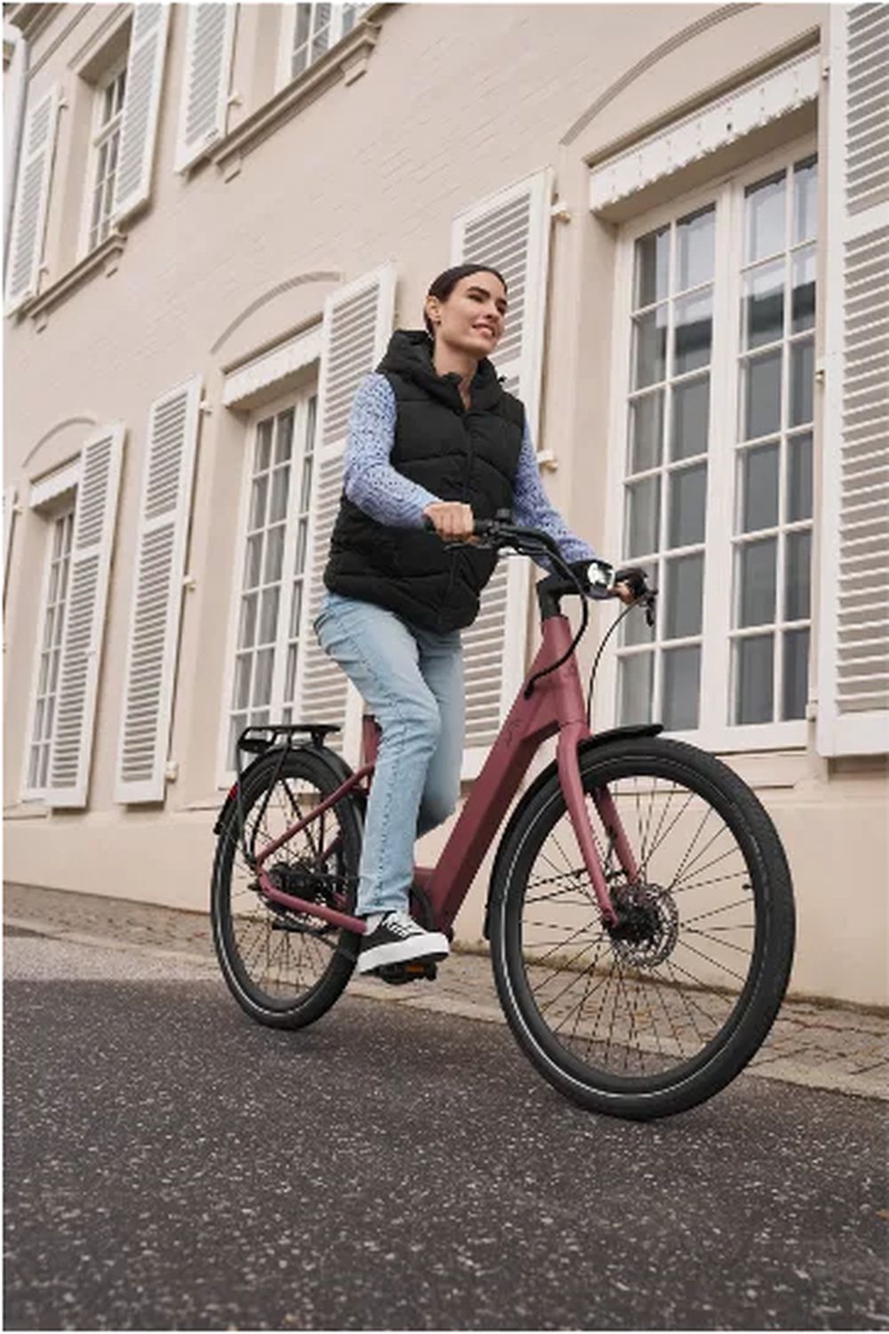 Mujer circulando en una bicicleta eléctrica CRIVIT Urban Y.3 rosa junto a un edificio de fachada clara.