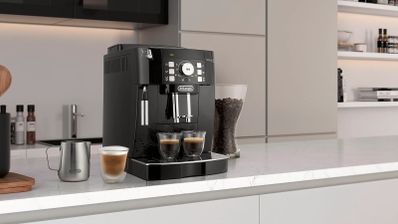 Cafetera De’Longhi Magnifica S negra sobre una encimera de cocina, con dos cafés bajo el dispensador y una jarra de leche al lado.