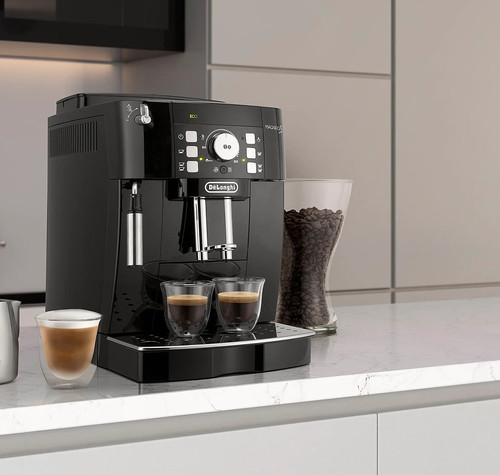 Cafetera De’Longhi Magnifica S negra sobre una encimera de cocina, con dos cafés bajo el dispensador y una jarra de leche al lado.