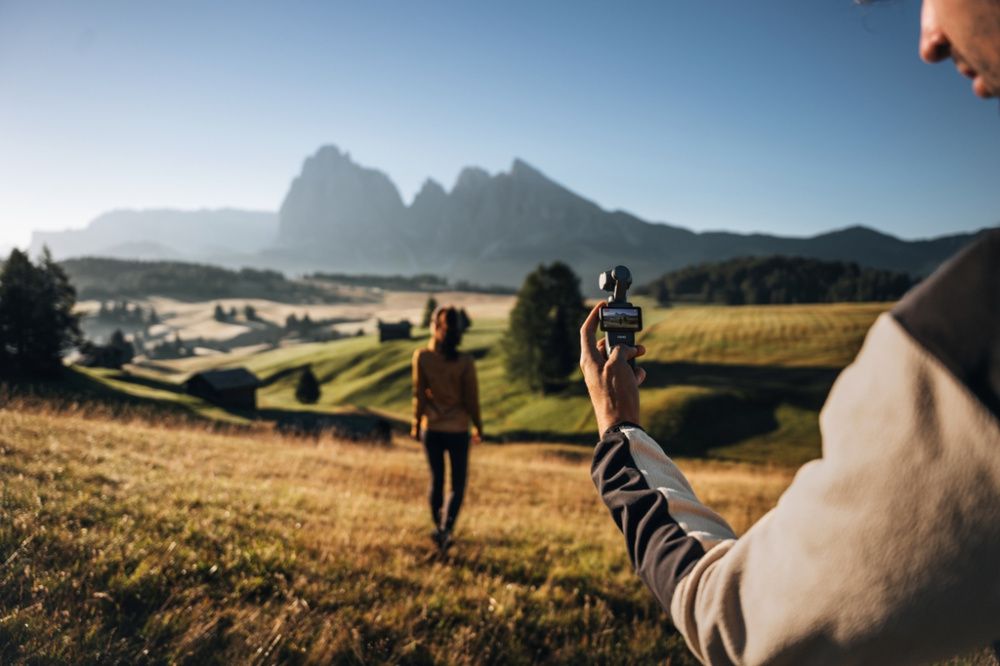 Persona utilizando una DJI Osmo Pocket 3 para grabar a una mujer caminando por un paisaje de montaña.