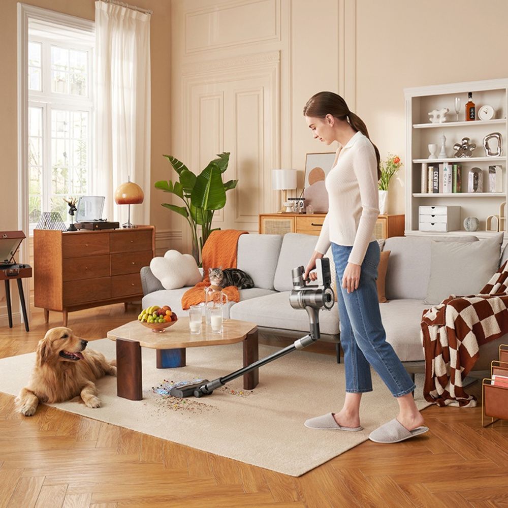 Una mujer utiliza la Dreame R20 para aspirar bajo una mesa de centro, con un perro y un gato en el salón.