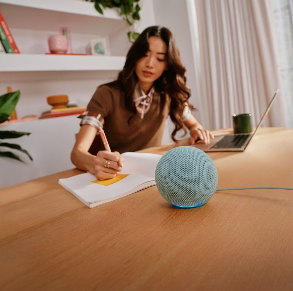 Echo Dot azul sobre un escritorio mientras una mujer escribe en un cuaderno junto a un portátil.