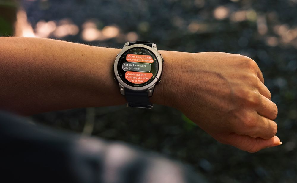 Garmin Fēnix E en la muñeca de una persona, con una conversación en pantalla y vegetación de fondo.