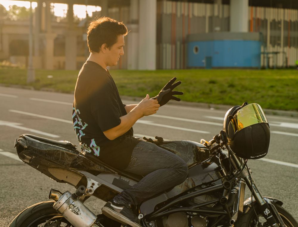 Hombre sentado sobre una motocicleta mientras se coloca unos guantes negros, con el casco apoyado delante.