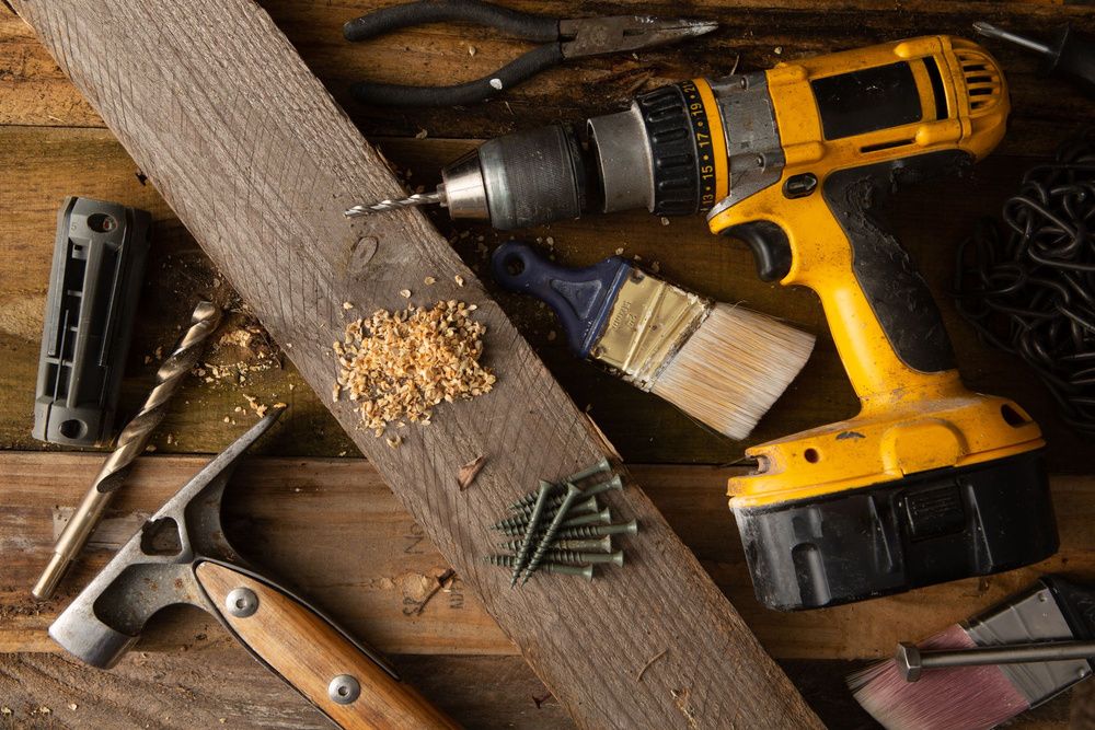 Taladro amarillo junto a tornillos, una brocha y otras herramientas sobre una superficie de madera.