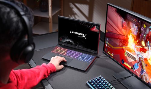 Una persona utiliza un portátil HyperX OMEN 15 con teclado iluminado junto a un monitor gaming en un escritorio.