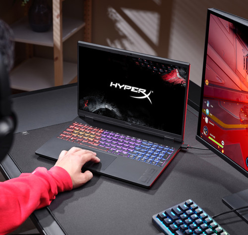 Una persona utiliza un portátil HyperX OMEN 15 con teclado iluminado junto a un monitor gaming en un escritorio.