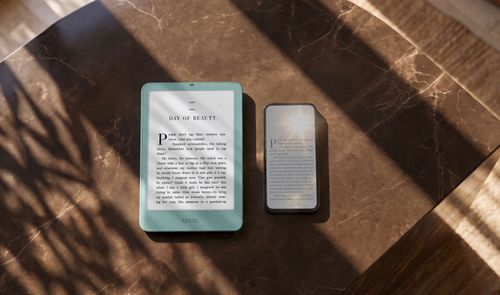 Kindle Paperwhite verde junto a un teléfono móvil sobre una mesa de mármol iluminada por el sol.