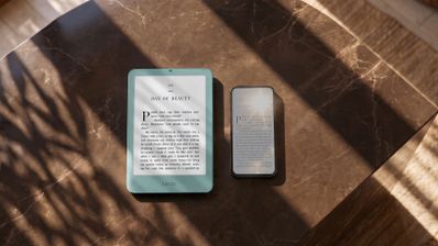 Kindle Paperwhite verde junto a un teléfono móvil sobre una mesa de mármol iluminada por el sol.
