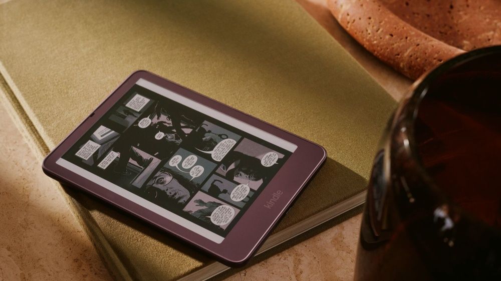 Kindle Colorsoft en tono morado mostrando un cómic, apoyado sobre un libro junto a una taza.