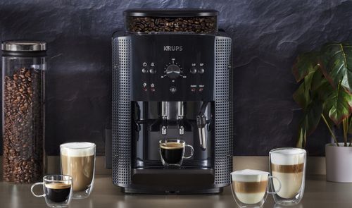 Cafetera KRUPS Roma negra sobre una encimera, acompañada de cafés, bebidas con leche, un recipiente de granos y una planta.