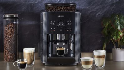 Cafetera KRUPS Roma negra sobre una encimera, acompañada de cafés, bebidas con leche, un recipiente de granos y una planta.