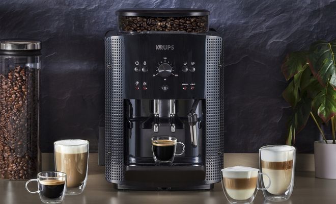 Cafetera KRUPS Roma negra sobre una encimera, acompañada de cafés, bebidas con leche, un recipiente de granos y una planta.