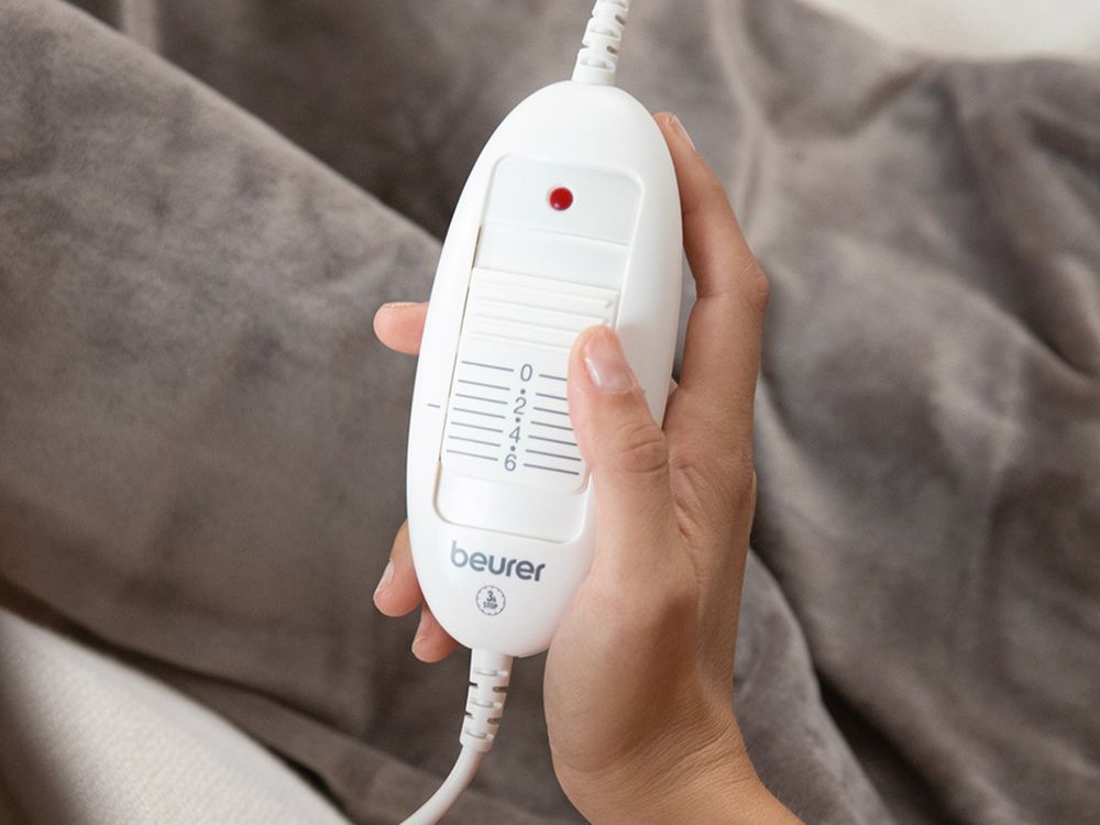 Mano sujetando el mando blanco de una manta eléctrica Beurer sobre un tejido gris.