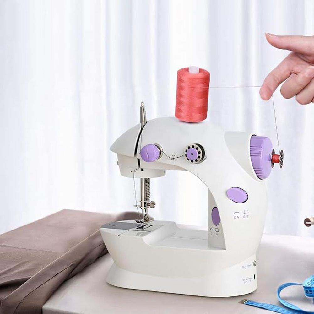Máquina de coser mini blanca y violeta sobre una mesa, con una bobina de hilo rojo y tela a su lado.